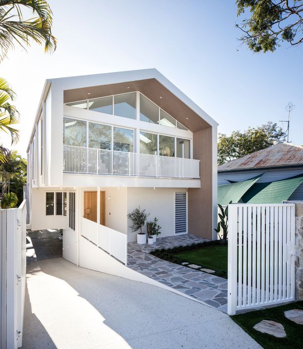 Contemporary two-storey residential home viewed from the front, featuring a crisp white facade, extensive upper-level glazing, a projecting gable-like roof form with a timber-lined soffit, white vertical metal balustrades and fencing, and a landscaped entry path of irregular stone paving. A covered carport or access area is visible beneath the upper floor, with neighbouring homes and tropical vegetation surrounding the property.