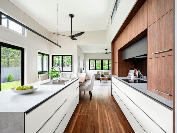 Bright open-plan contemporary kitchen and dining/living space with a long white island, integrated stainless-steel sink and tap, white handleless cabinetry, dark stone-look benchtops, and a full-height warm timber built-in wall with concealed storage. Timber flooring extends through the connected dining and living area, with extensive black-framed glazing, clerestory windows, and ceiling fans creating an airy, spacious interior.