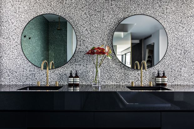 A sophisticated bathroom vanity with a long dark stone countertop and black built-in cabinetry, featuring two round mirrors, twin gold-toned faucets and vessel sinks, and a patterned grey-and-white feature wall. The mirrors reflect a dark tiled shower area and adjacent modern interior. A small floral arrangement and coordinated dispensers add decorative detail.