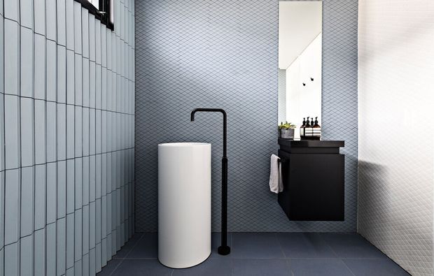 Contemporary minimalist bathroom featuring a freestanding white cylindrical basin, matte black wall-mounted vanity, tall mirror, black floor-mounted tap, and geometric textured wall and floor tiles. A vertical tiled screen separates the basin area from the rest of the space.