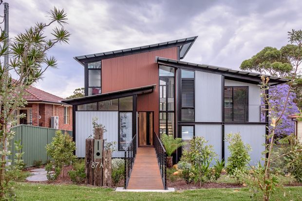 Street-facing view of a compact contemporary multi-level residence with an asymmetrical pitched roof, projecting roof planes, dark metal-framed glazing, and corrugated metal wall cladding in contrasting red and light grey tones. A timber walkway leads through landscaped planting to the recessed front entry. Neighbouring homes and established vegetation are visible around the property.