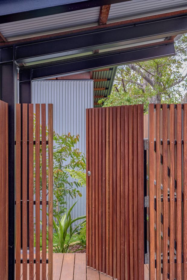 Close-up view of a contemporary exterior entry or courtyard gate, featuring vertical reddish-brown timber slats, a solid timber pedestrian gate, dark metal framing, corrugated metal roofing and cladding, and dense planting visible behind the fence.