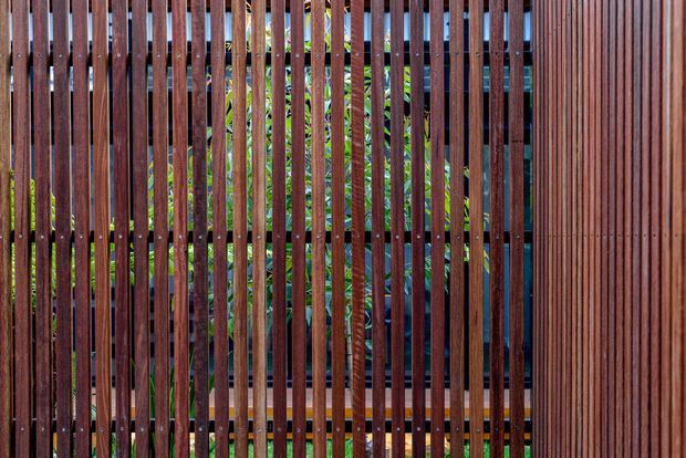 Close-up view of a contemporary exterior screen or fence composed of closely spaced vertical timber slats in a rich reddish-brown finish. Green planting and parts of a dark building structure are visible through the gaps. The repetitive linear pattern creates privacy while allowing filtered light and views.