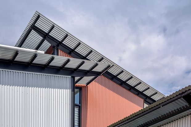 Close-up exterior view of a contemporary building with corrugated metal cladding in light grey and muted terracotta, dark structural framing, and prominent projecting slatted metal awnings or canopies. The angular roofline and layered canopy geometry are set against an overcast sky.