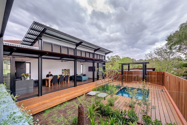 Exterior view of a contemporary multi-level home with a covered timber deck, open-plan dining and living areas visible through extensive glazing, dark metal framing, and a timber-fenced backyard with a small swimming pool and deck. The building has a low-pitched corrugated roof and a broad covered outdoor entertaining area.