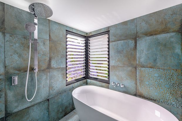 A modern bathroom featuring a freestanding white bathtub positioned beneath a corner window with louvred shutters. The walls are clad in large blue-green textured patterned tiles, complemented by a wall-mounted brushed metal rain shower and handheld shower set. The space has a clean, contemporary feel with natural light entering through the windows.