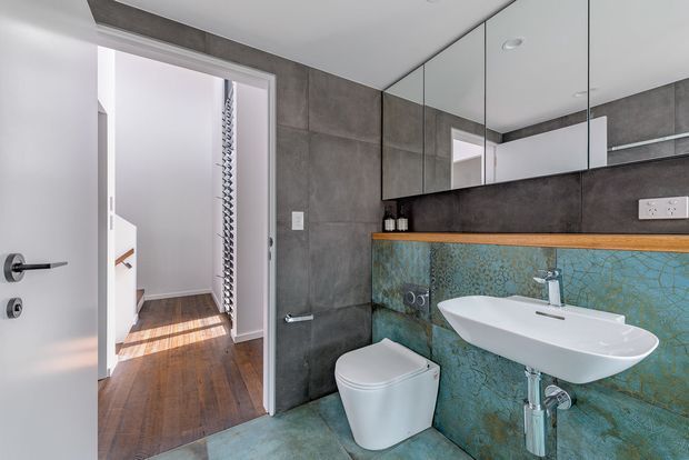 Contemporary bathroom with large grey wall tiles, a freestanding-style white basin, wall-mounted toilet, mirrored cabinetry, and a turquoise patterned feature wall behind the vanity. The open doorway reveals a timber-floored hallway and stair area.