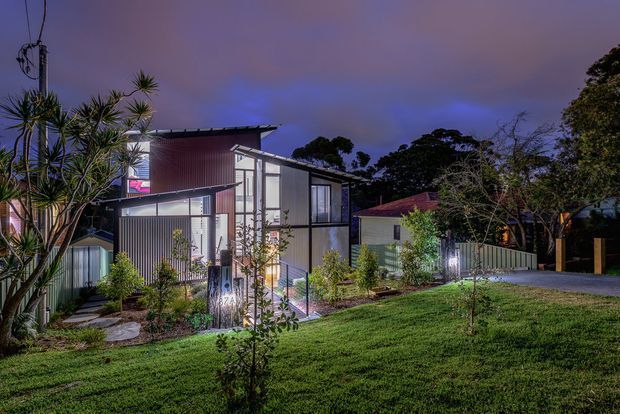 Dusk exterior view of a contemporary multi-level residence set within a landscaped suburban garden. The home features dark metal cladding, extensive floor-to-ceiling glazing, a projecting upper volume, and illuminated interiors. Mature trees, lawn, planted garden beds, and a neighbouring pitched-roof structure are visible around the building.