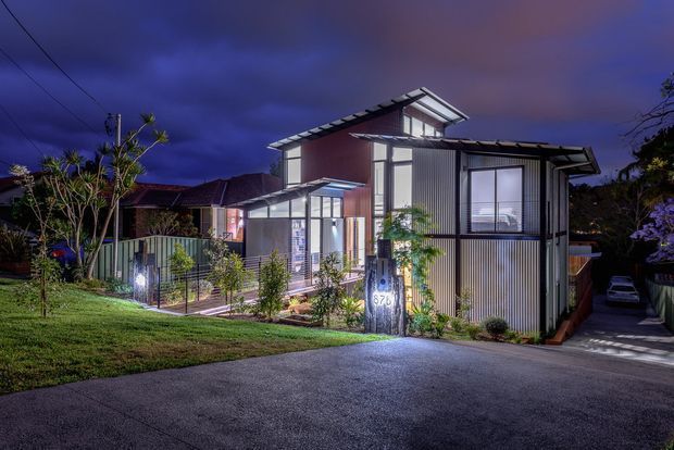 Twilight exterior view of a contemporary multi-level residential house on a suburban site. The home features strong angular roof forms, extensive glazing, vertically ribbed light-coloured cladding, dark structural framing, and illuminated landscaped entry paths. A driveway and neighbouring residential context are visible.