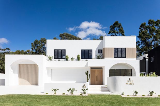 A bright contemporary two-storey white rendered house viewed from the front, featuring strong geometric volumes, rounded arched openings, a large integrated garage, timber-look vertical screening, dark-framed windows, a timber entry door, and minimalist landscaped planting.