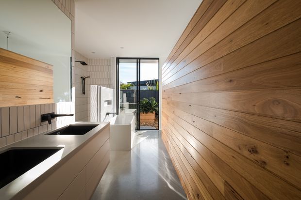 A bright contemporary bathroom with a long, narrow layout. The room features a double vanity with pale cabinetry and dark inset basins, a freestanding white bathtub, tiled walls, a full-height timber feature wall, polished concrete flooring, and black-framed glazing opening toward a small courtyard. Natural light enters through the glass door and window.