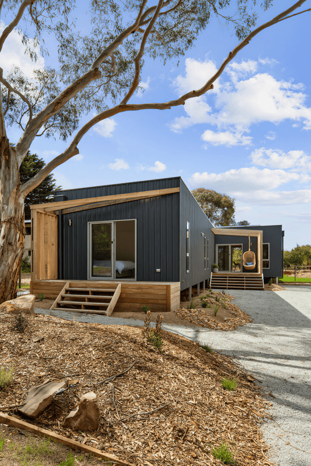 Exterior view of a contemporary compact modular or prefabricated building composed of two dark corrugated-clad rectangular volumes connected by a gravel approach. The structures feature timber-framed entries, raised timber decks and steps, large glazed doors, and low-pitched roof forms, set beneath a mature eucalyptus tree in a landscaped natural site.