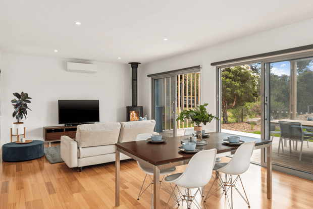 Bright open-plan living and dining area with warm timber flooring, white walls, a cream sofa, dark wood dining table, and white sculptural dining chairs. Large sliding glazed doors connect the room to an outdoor deck or patio, with a compact fireplace and television along the living-room wall. The space has a clean, contemporary residential design with abundant natural light.