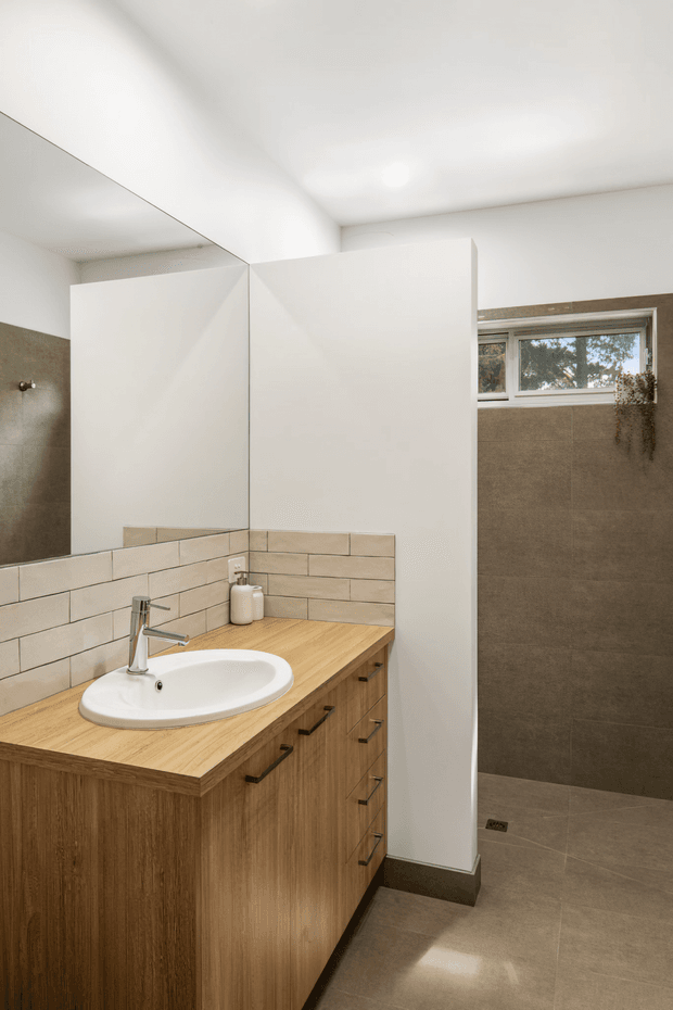A bright contemporary bathroom featuring a timber-look vanity with a light wood benchtop, white vessel basin and chrome tapware. A large wall mirror sits above the vanity, while the shower area is separated by a white privacy partition and finished with large taupe-grey wall and floor tiles. A narrow horizontal window provides natural light, with a small hanging plant adding subtle greenery.