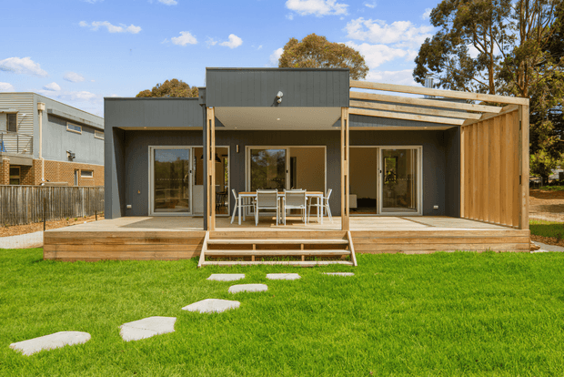 A contemporary single-storey home viewed from the rear, with dark grey cladding, extensive timber decking, a large timber-framed covered outdoor area, and full-height glazed doors opening onto a dining and living space. A manicured lawn with stepping stones occupies the foreground, with neighbouring homes and mature trees in the background.