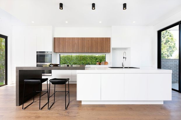 Bright contemporary kitchen with white full-height cabinetry, a large white island with integrated sink, dark stone benchtops, timber flooring, and a warm timber feature cabinet above the rear bench. Black-framed glazing, black pendant-style ceiling fixtures, and two black stools provide contrast. The kitchen opens toward greenery and an exterior brick wall visible through the windows.