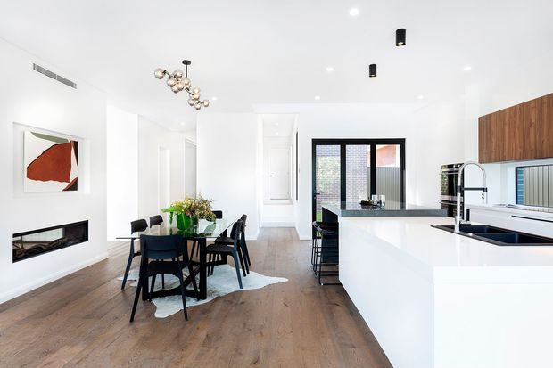 Bright, open-plan contemporary kitchen and dining area with white cabinetry and walls, a large white island with dark benchtop, integrated appliances, timber flooring, black-framed glazing, and a dining table beneath a sculptural pendant light. A living area with fireplace is visible at the left.