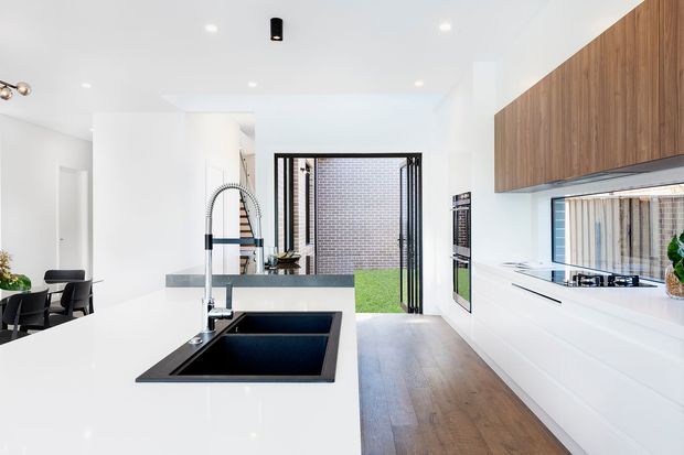Bright contemporary open-plan kitchen featuring a large white island with integrated black double sink and gooseneck mixer, white lower cabinetry, and warm timber overhead cabinetry. A dark stone-look benchtop runs along the rear cooking zone, with integrated appliances and a splashback window. Timber flooring extends toward a glazed rear opening, while a dining area is visible to the left. The space has clean white walls, recessed downlights, and strong indoor-outdoor connection.