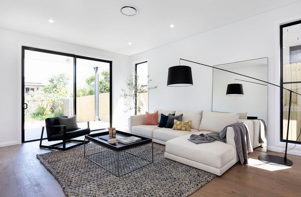 Bright contemporary living room with white walls and ceiling, warm timber flooring, black-framed glazing, a large pale sectional sofa, black lounge chair, low black coffee table, textured rug, oversized black pendant lighting, and a large wall mirror. Sliding glass doors open to an outdoor area, while a staircase is visible through the glazing at right.