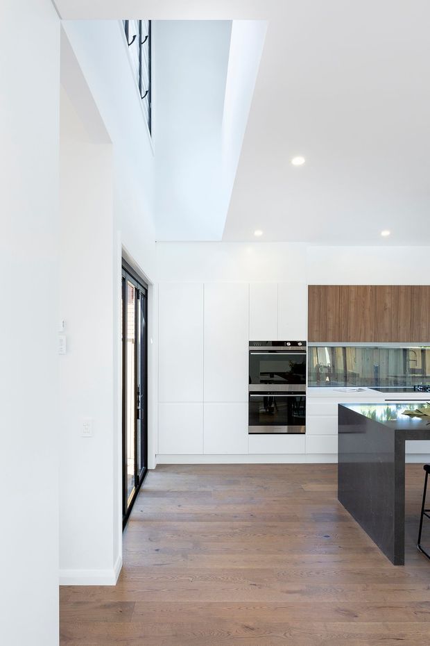 Bright contemporary kitchen interior with white full-height cabinetry, integrated black oven, warm timber upper cabinetry, a dark stone island, and wide-plank timber flooring. Black-framed glazing opens along the left side, while recessed ceiling lights and a large ceiling void/skylight-like opening enhance the spacious feel.