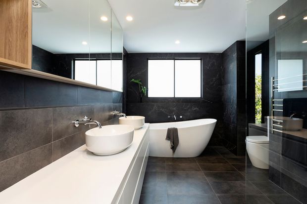 Contemporary luxury bathroom with a freestanding white bathtub, double vessel basins on a long white vanity, dark stone-look wall and floor finishes, large mirrored cabinetry, and black-framed windows. The room has a clean, high-contrast monochromatic palette with integrated ceiling lighting and a partially visible shower and toilet area.
