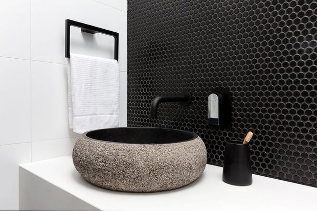Close-up of a contemporary bathroom vanity featuring a round, rough-textured stone vessel basin on a white benchtop. The wall behind combines large white tiles with a dark circular mosaic tile feature, complemented by matte black wall-mounted tapware, a black towel rail, and a black toothbrush holder.