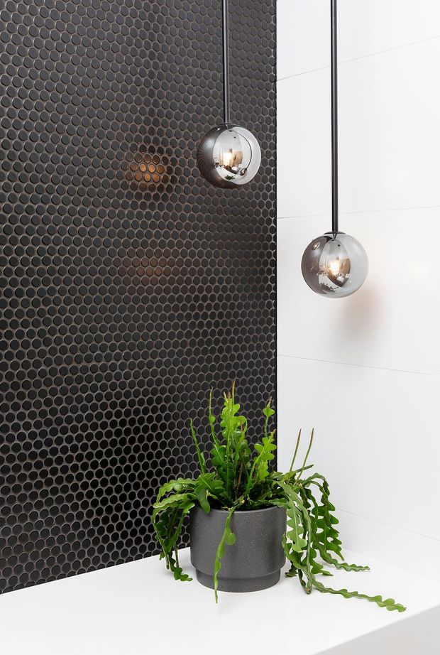 Close-up interior detail featuring a dark charcoal circular mosaic tile feature wall beside large white wall panels. Two chrome spherical pendant lights hang in front of the wall, with a dark textured planter holding lush green foliage on a white ledge or benchtop. The composition is crisp, contemporary, and high contrast.