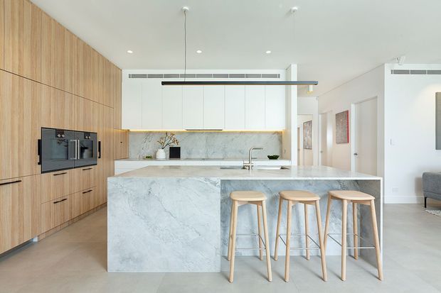 Bright contemporary kitchen with a large marble-look island and three timber bar stools. Full-height light timber cabinetry with integrated appliances lines the left wall, while a pale stone splashback, white cabinetry, and minimalist pendant lighting create a clean open-plan interior.