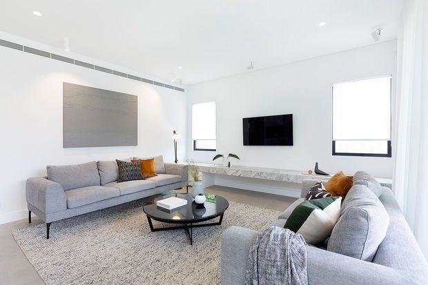 Bright contemporary living room with white walls and ceiling, large windows, two light-grey sofas, a textured neutral rug, a black oval coffee table, and a long pale stone or concrete-look built-in media console. Minimal decor and recessed lighting create an airy, refined interior.