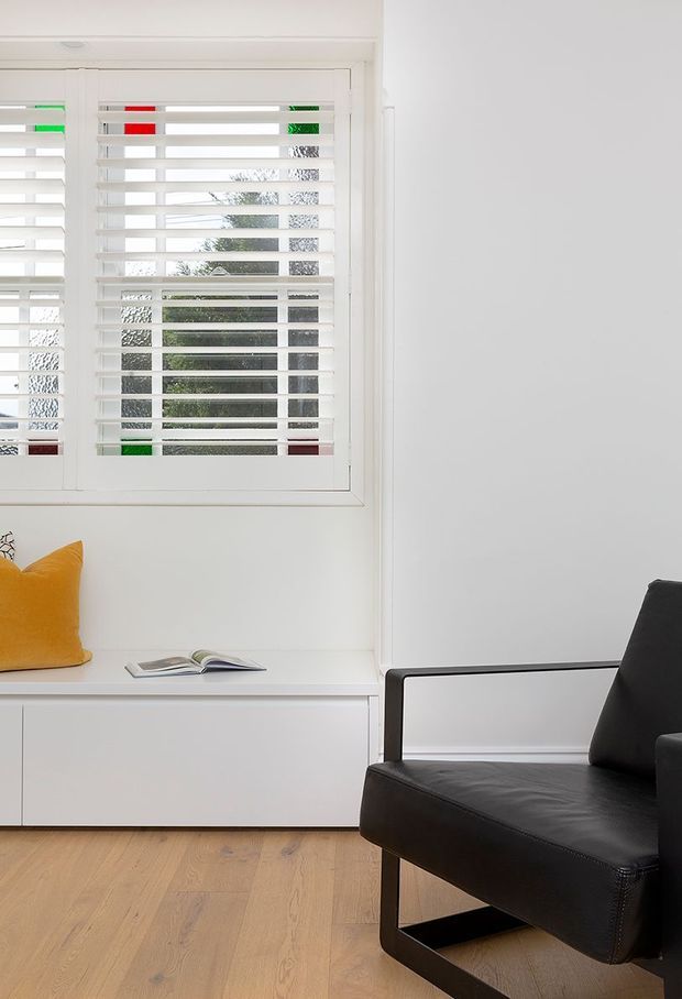 Bright, minimalist interior corner featuring a large white-framed window with horizontal plantation shutters, a built-in white window seat or low cabinet, and a black leather armchair. The room has white walls and light timber flooring, with a mustard-yellow cushion adding a small accent of colour. Greenery and neighbouring structures are visible through the shutters.