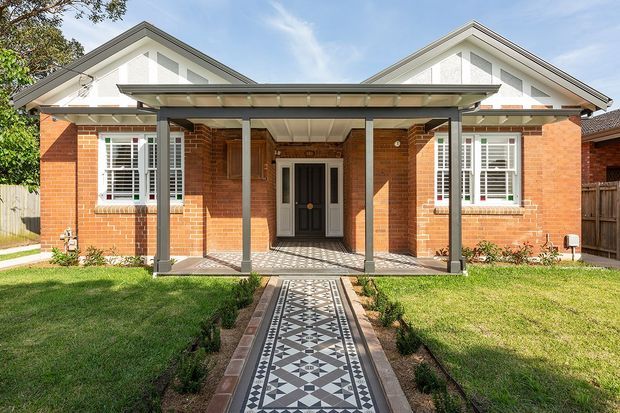 Front exterior view of a symmetrical brick residence with two white gabled roof forms, a broad covered entry porch supported by slim dark posts, plantation-shuttered windows, and a decorative geometric tiled path leading to the central front door. The facade is brightly lit and set behind a neat lawn.