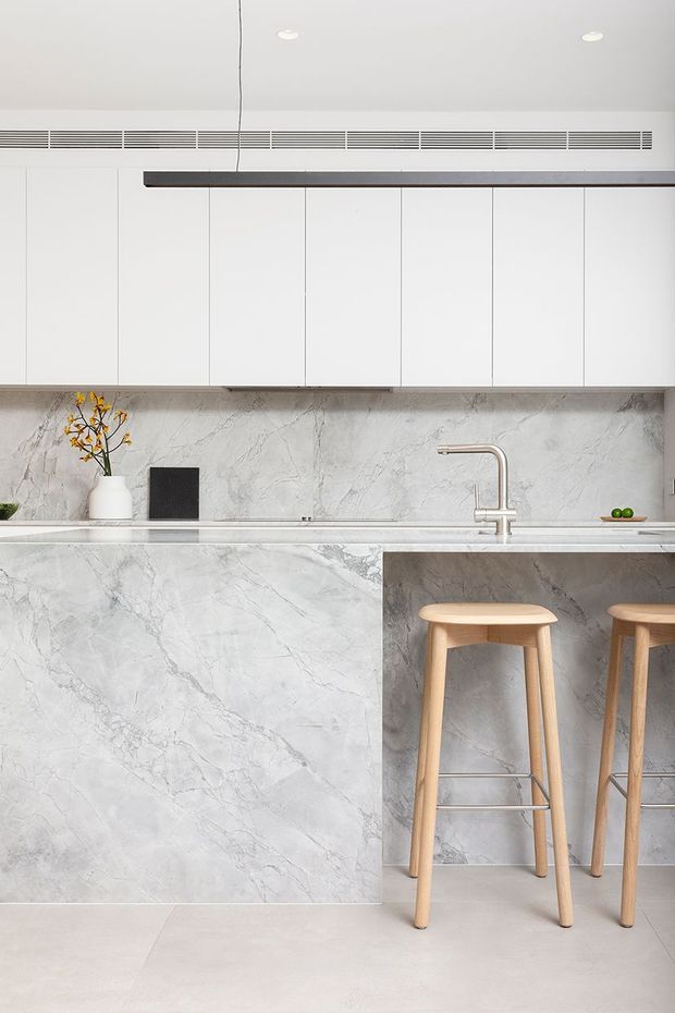 A minimalist contemporary kitchen featuring a long island clad in pale grey veined stone, matching stone backsplash, white handleless overhead cabinetry, and a slim dark linear pendant light. A brushed metal tap and sink sit at the island, with two light timber bar stools tucked beneath the overhang. The space has a clean, refined, neutral palette and integrated detailing.