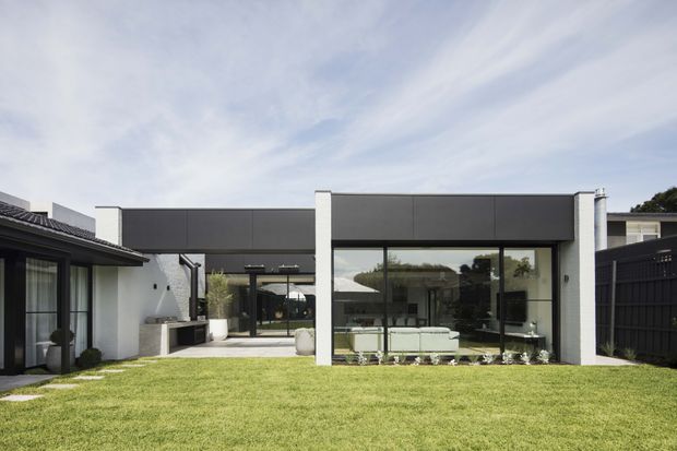 Exterior view of a low, contemporary residential extension arranged around a broad grass courtyard. The building has a flat roof, dark charcoal cladding bands, white rendered structural walls, extensive floor-to-ceiling glazing, and a covered outdoor living area. A furnished living room is visible through the glazing, with an older tiled-roof wing adjoining on the left.