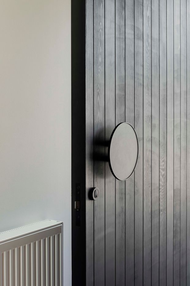 Close-up interior view of a contemporary vertically slatted timber door with a dark frame, circular inset or feature panel, and visible lock hardware. A white wall and panel radiator sit beside the door. The image is tightly framed around the architectural door detail.