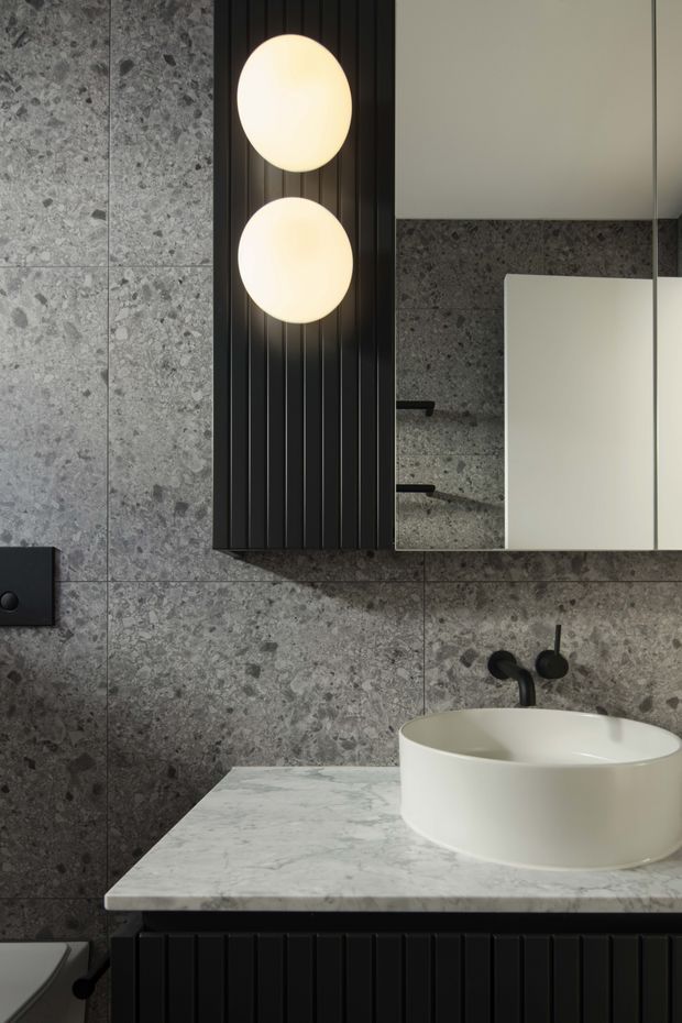 A dark, refined bathroom vanity vignette featuring large grey stone-look wall tiles, a white marble-look benchtop, a round white vessel basin, matte black tapware, and black fluted vertical-panel joinery. A mirrored cabinet and two glowing spherical wall lights add contrast and a contemporary decorative focal point.