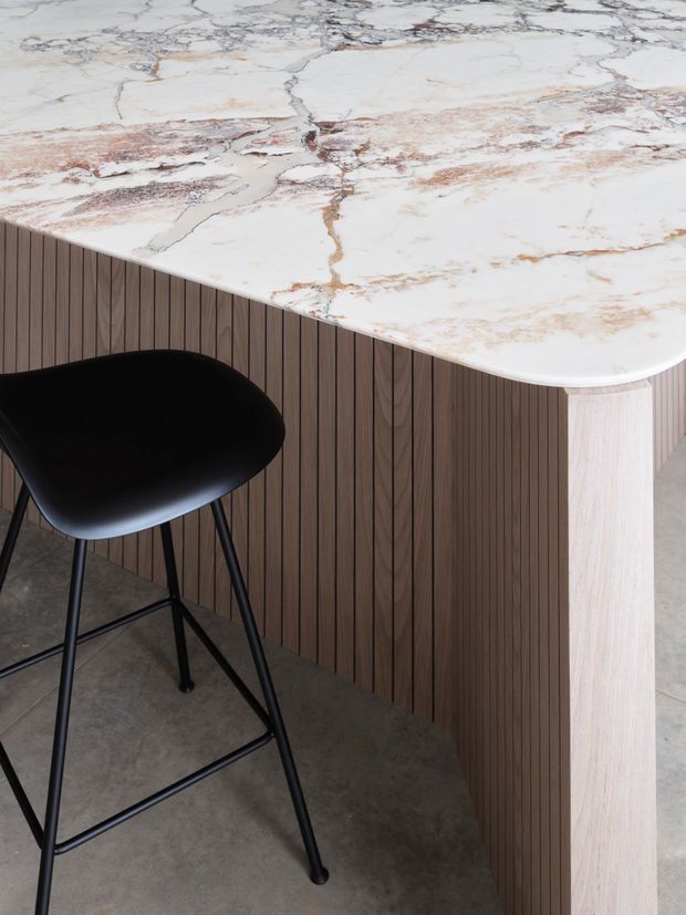 Close-up of a contemporary kitchen island or counter with a rounded white marble-look stone benchtop featuring prominent grey and warm brown veining. The island has vertically ribbed dark timber panelling and a pale timber end support, paired with a black bar stool. A grey polished concrete floor is visible below.