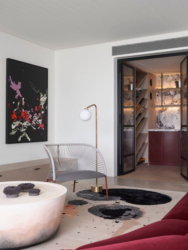Refined contemporary living room with a sculptural white coffee table, wire-frame lounge chair, brass floor lamp, burgundy sofa, patterned area rug, and large abstract artwork. A dark-framed opening leads to a compact walk-in pantry or bar area with timber shelving, marble-patterned wall surfaces, and burgundy cabinetry. The room has a white paneled ceiling and a restrained palette of white, black, burgundy, timber, and brass.