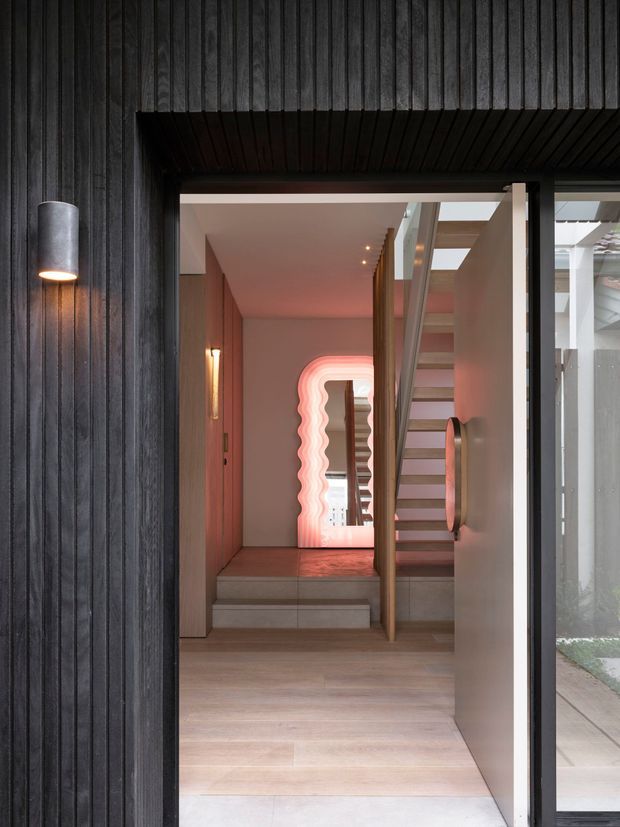 A modern residential entry viewed from outside through an open doorway. The facade is clad in dark, vertically ribbed timber, with a cylindrical wall light beside the entrance. Inside, a warm-toned timber or wood-look floor leads to a short stair and open stairway with slim timber and metal elements. Pink ambient lighting, skylights, and a sculptural wavy illuminated opening create a dramatic contemporary feature.