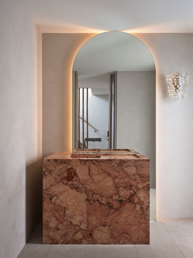 A refined bathroom vanity featuring a substantial reddish-brown veined natural stone cabinet and integrated double basins. A large arched mirror with concealed warm backlighting dominates the wall, complemented by a sculptural glass wall sconce. The space has pale plastered walls, light tiled flooring, and a stairway visible in the mirror reflection.