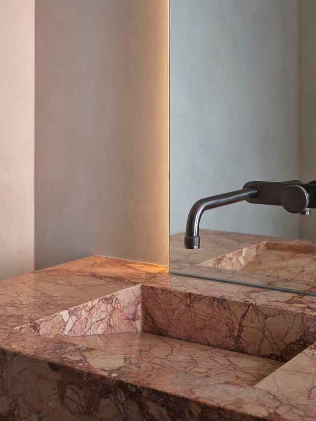 Close-up of a refined bathroom vanity featuring a carved pink and burgundy natural stone basin and countertop, with a dark bronze wall-mounted tap reflected in a large mirror. Warm concealed lighting highlights the stone surface and creates a luxurious, sculptural atmosphere.