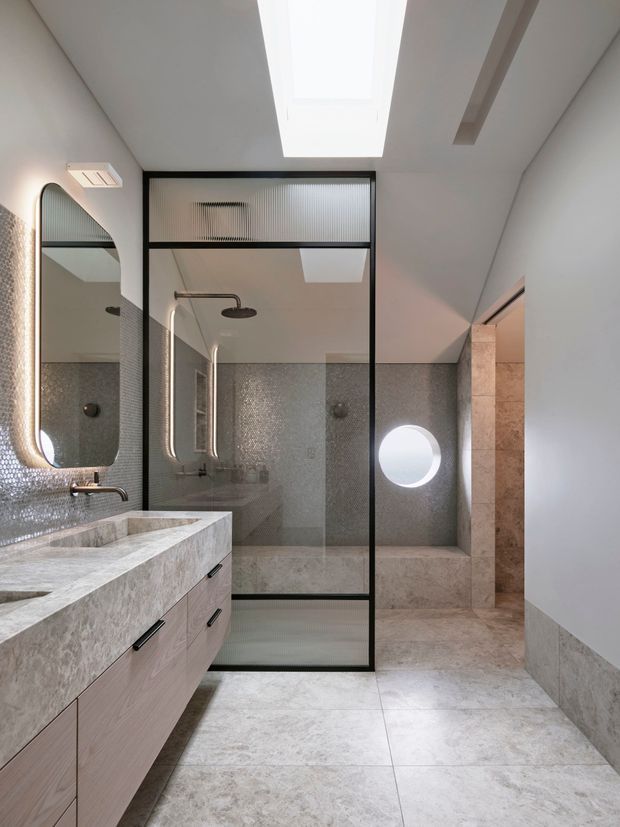 Contemporary bathroom featuring a long stone vanity with integrated basin, pale wood cabinetry, large-format stone-look floor and wall tiles, a glass shower enclosure with black metal framing and fluted glass panels, an arched illuminated mirror, wall-mounted fixtures, skylights, and a circular window. The space is bright, refined, and softly neutral.