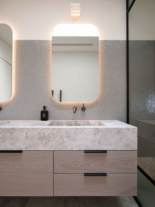 Contemporary bathroom vanity with a large integrated pale stone trough basin, timber-look drawers, brushed metal wall-mounted tapware, and softly backlit rounded-rectangle mirrors. Small-format mosaic tiles cover the wall, with a dark framed glazed shower enclosure at right.