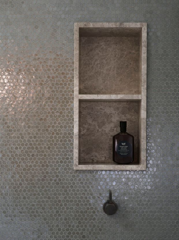 Close-up of a bathroom shower wall clad in small grey hexagonal tiles, featuring a vertically stacked recessed niche finished in mottled brown-grey stone and holding a dark soap bottle. A dark circular shower control is visible below.