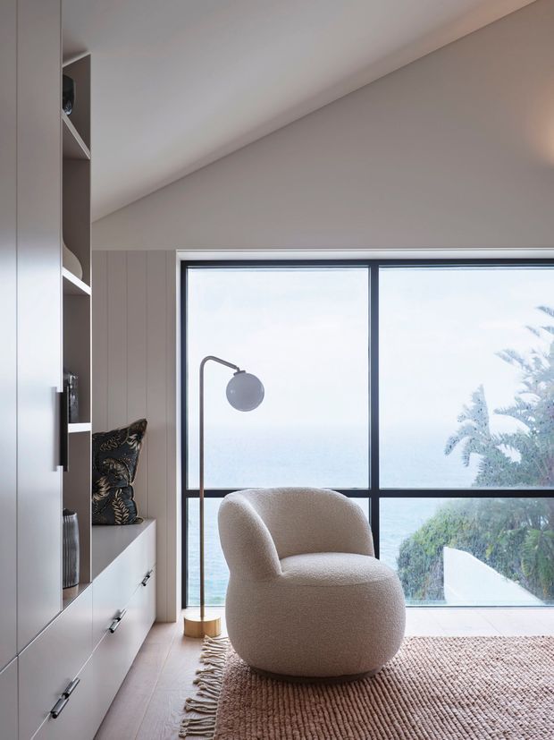 A softly styled reading nook beside a large black-framed window, featuring a curved upholstered lounge chair, slim floor lamp, built-in white cabinetry and shelving, and a textured neutral rug. The window overlooks a bright coastal landscape, while vertical wall panelling and a sloped ceiling add architectural character.