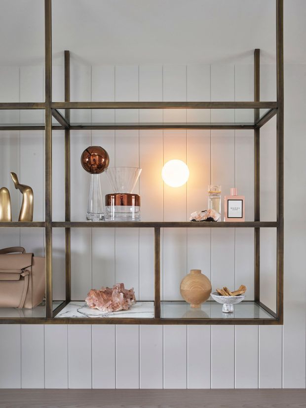 A close-up interior view of a floor-to-ceiling open display shelving unit with dark bronze-toned metal framing, white vertically panelled backing, and marble shelves. The shelves are styled with glassware, decorative vessels, perfume bottles, a handbag, sculptural objects, and a glowing spherical lamp. The composition has a refined, curated contemporary feel with warm metallic and blush accents.
