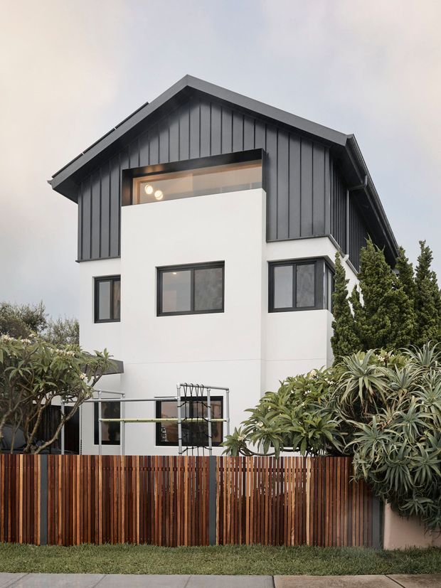 A contemporary multi-storey residential house viewed from the street, with a crisp white rendered facade, dark vertical metal cladding and a steep gabled roof. Black-framed windows and a projecting upper-level volume create strong geometric contrast. A timber slat boundary fence, planted garden, and mature shrubs occupy the foreground.