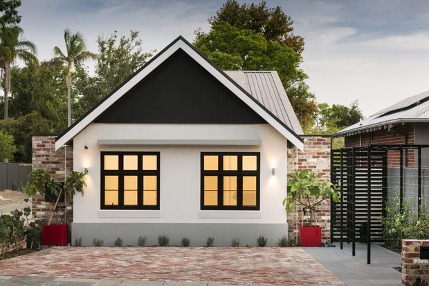 Front exterior view of a compact gabled building with white rendered walls, a dark triangular gable accent, black-framed multi-pane windows, a corrugated metal roof, exposed brick side walls, and a brick-paved forecourt. Tropical planting and a slatted black privacy screen frame the entrance area.