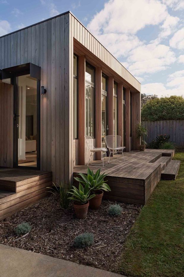 A compact contemporary single-storey pavilion or extension clad in vertical timber boards, with a simple flat roof and a deep recessed veranda. Tall timber-framed glazing lines the facade, opening onto a raised timber deck with integrated bench seating and a rectangular recessed feature. The garden includes mulched planting beds, potted plants, lawn, and a timber boundary fence. A glazed entry door reveals part of a light-coloured interior.
