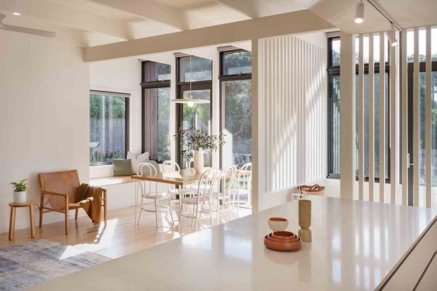 Bright open-plan interior with a contemporary kitchen island in the foreground and a dining area beside expansive dark-framed windows. White surfaces, pale timber flooring, sculptural dining chairs, a small lounge nook, and vertical timber screening create an airy, light-filled space. Exposed ceiling beams and a partially visible courtyard or garden beyond the glazing add architectural interest.