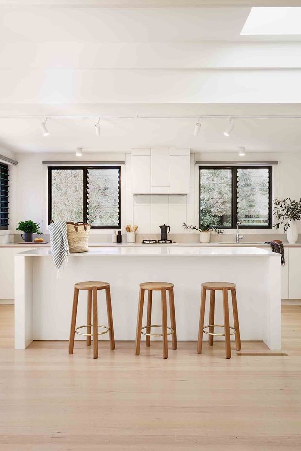 Bright, contemporary white kitchen with a large island and three timber stools. The room features white flat-panel cabinetry, a tiled splashback, pale timber flooring, dark-framed windows with plantation shutters, a skylight, and track lighting. Minimal styling includes potted plants, woven baskets, and simple kitchen accessories.